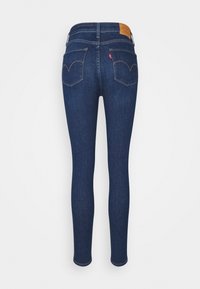 Jean skinny bleu foncé avec des poches arrière, des passants pour ceinture et une étiquette de marque en cuir marron sur la ceinture.