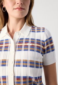 Polo blanc avec un motif à carreaux multicolores comportant du bleu, du jaune et du rouge. Manches courtes et col classique avec fermeture à bouton.