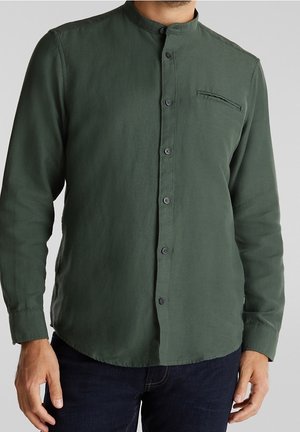 Homme portant une chemise à col mandarin vert foncé à manches longues et à boutons sur le devant, associée à un jean bleu foncé.