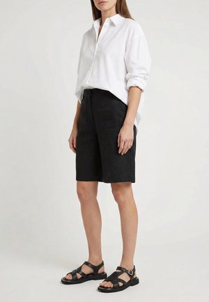 Kvinde iført en hvid langærmet skjorte, sorte shorts til knæet og sorte åbne sandaler, stående mod en ensfarvet baggrund.