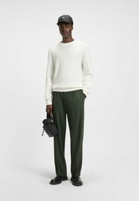 Weißer strukturierter Pullover, olivgrüne Hose, schwarze Mütze und eine kleine schwarze Tasche. Die Schuhe sind flach und schwarz, mit einem dezenten Glanz.