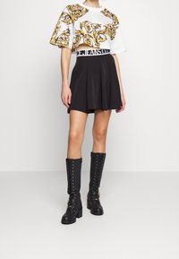 Bílá zkrácená košile s pozlaceným zdobením, kombinovaná s černou rozšířenou sukní. Vysoké černé boty s tkaničkami a zlatými akcenty.