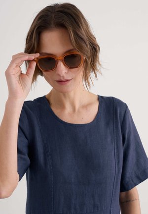 Woman with short wavy brown hair adjusting amber-framed sunglasses, wearing a navy blue short-sleeve linen top against a plain background.