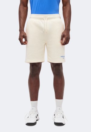 Άνδρας που φορά λευκά σορτς με κορδόνι και μικρό μπλε κείμενο στο αριστερό πόδι, λευκές κάλτσες, λευκά και μαύρα αθλητικά παπούτσια, στέκεται μπροστά σε απλό φόντο.