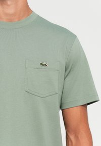 Lichtgroene t-shirt met korte mouwen en een borstzak, voorzien van een klein geborduurd krokodillenlogo.