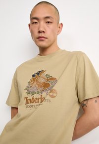 T-shirt en coton beige présentant un graphique coloré de bottes parmi des motifs de plantes abstraits, avec le texte "Timberland" en marron en dessous.
