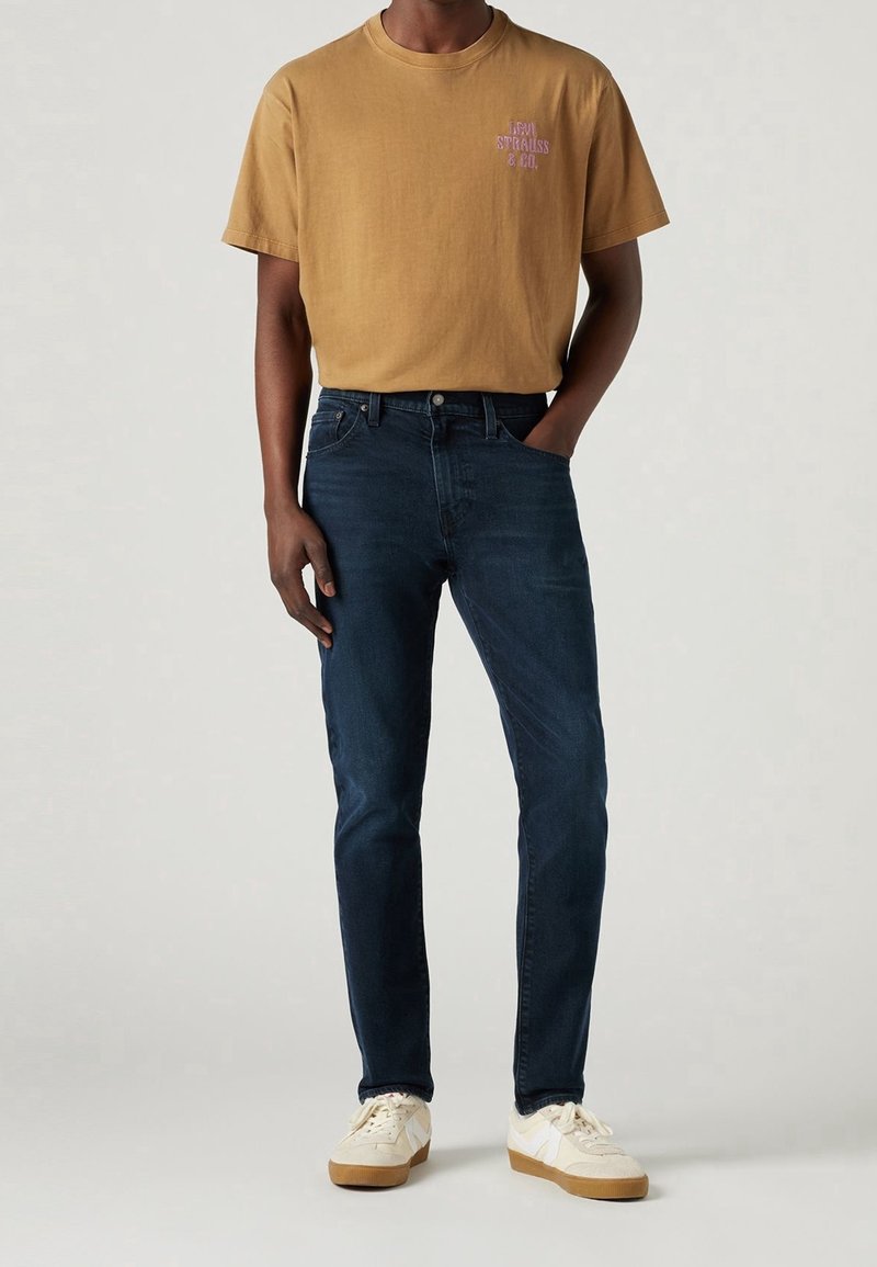 Mörkblå denimjeans i kombination med en senapsgul kortärmad T-shirt med lila text, bärs med beige sneakers.