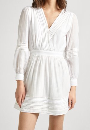 Vrouw in een witte jurk met lange mouwen, een V-hals, een geraffineerde taille, verticale plooien en kantafwerking op de mouwen en de zoom.