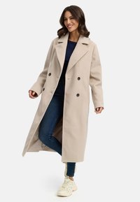 Beige wollen blend jas met een dubbelzijdig ontwerp, grote revers en twee voorzakken, gedragen over een blauwe jeans en lichte sneakers.