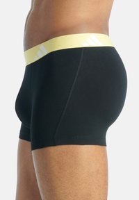 Svarta boxershorts i slät tyg, med en gul midja som har vita ränder och logotyp. Sidovy visar design som sitter tajt.