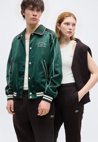 Νέος άνδρας με πράσινο μπουφάν του κολεγίου και σκούρο παντελόνι, νέα γυναίκα με λευκή φανέλα χωρίς μανίκια και μαύρο μπουφάν χωρίς μανίκια περασμένο στους ώμους, και οι δύο όρθιοι.