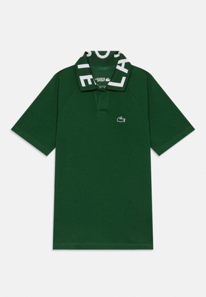Polo shirt à manches courtes vert foncé avec un logo Lacoste blanc audacieux sur le col et un emblème crocodile brodé sur la poitrine.