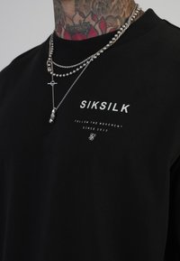 Camiseta de algodón negra con el logo de SIKSILK en blanco, cuello redondo, con cadenas plateadas superpuestas que presentan cuentas y un colgante.