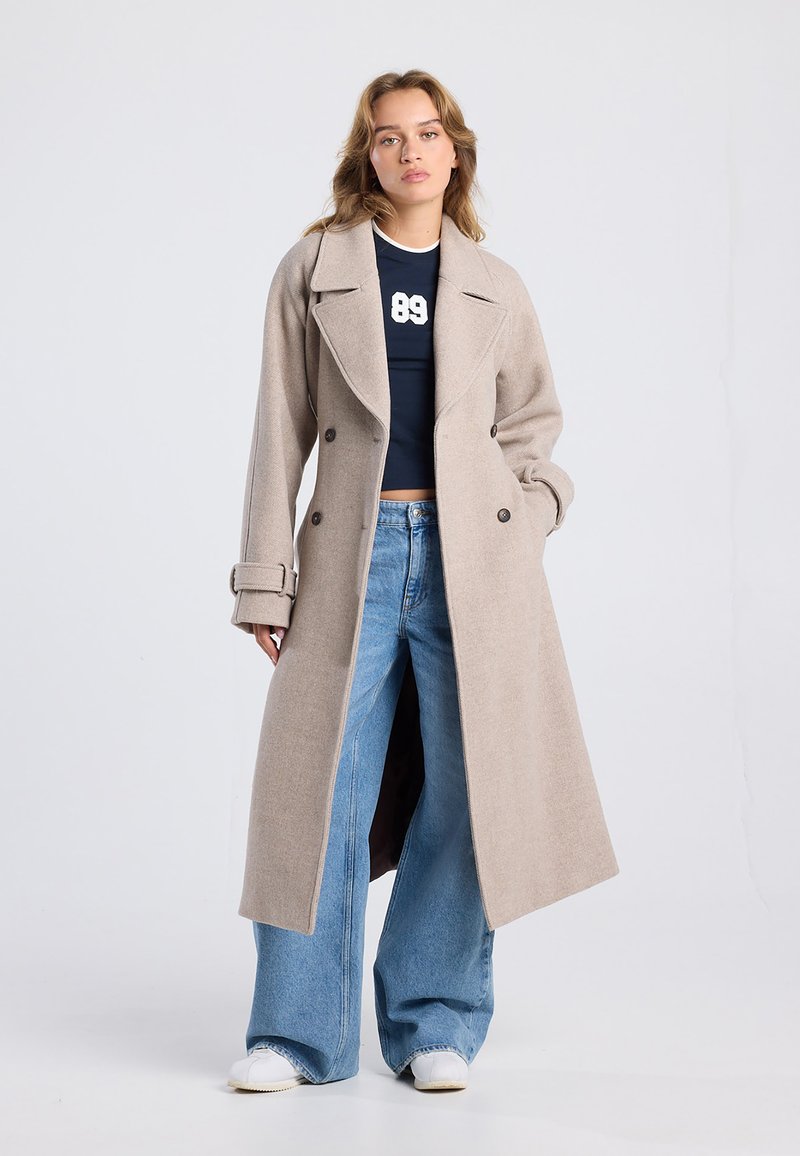 Abrigo de mezcla de lana beige con diseño de doble botonadura, solapas con muesca y puños enrollados, llevado sobre una camiseta azul marino y unos vaqueros de pierna ancha.