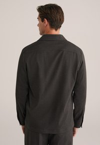 Hombre con cabello corto castaño que lleva una camisa de manga larga gris oscuro y pantalones a juego, de pie de espaldas frente a un fondo liso.