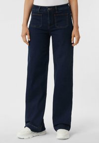 Jeans a gamba larga in denim blu scuro. Presentano due tasche anteriori e due tasche posteriori con cucitura a contrasto. Indossati con sneaker bianche.