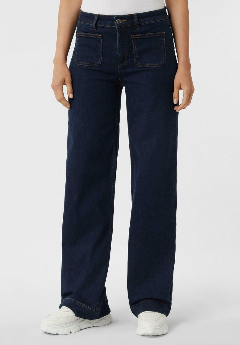 Jeans a gamba larga in denim blu scuro. Presentano due tasche anteriori e due tasche posteriori con cucitura a contrasto. Indossati con sneaker bianche.