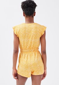 Combinaison courte jaune à motifs floraux, avec manches courtes à volants, taille élastiquée et coupe ample. Le tissu présente une finition subtilement texturée.