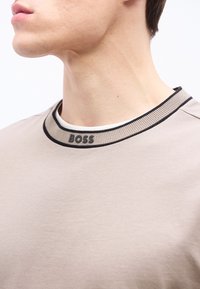 Κοντινό πλάνο του λαιμού και του κάτω μέρους του προσώπου ενός άνδρα που φοράει ένα μπεζ πουκάμισο με ριμπ κολάρο με μαύρη επένδυση και λογότυπο "BOSS".