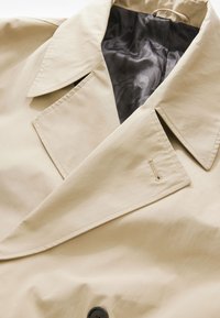 Abrigo trench beige con un amplio collar, dos grandes botones negros y un forro interior negro liso y brillante con patrones sutiles.