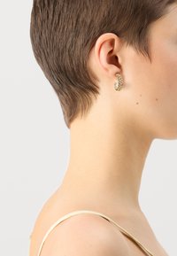 Brinco de argola em tom dourado com um design trançado, realçado por uma única pérola. Usado na orelha de um modelo com cabelo curto contra um fundo neutro.