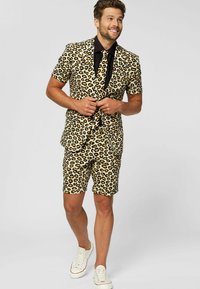 Leopardų rašto kostiumas, pagamintas iš audinio su smėlio spalvos pagrindu, turintis juodų ir rudų taškų, trumpomis rankovėmis, šortais ir juoda marškinėliais po. 