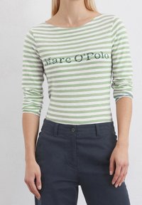 Vihreä-valkoinen raidallinen pitkähihainen paita, jossa on leveä kaula-aukko. Rintamuksessa on "Marc O'Polo" -logo vihreällä tekstillä. Paritettu tummien housujen kanssa.