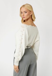 Maglione bianco a spalle scoperte in maglia con dettagli in pizzo e accenti di bottoni sulle maniche, abbinato a pantaloni grigi su misura.