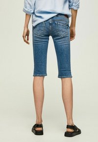 Pantalons capri en denim bleu moyen, avec une coupe ajustée, un ourlet roulé et des accents déchirés, agrémentés de poches arrière avec des détails de surpiqûre.