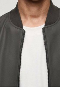 Chaqueta bomber gris con textura lisa, cuello y puños acanalados, y cierre de cremallera, usada sobre una camiseta blanca.