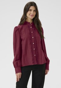 Bordowa bluzka z wysokim kołnierzem, zapinana na guziki, z koronkowym wykończeniem wzdłuż plisy i mankietów. Miękka tkanina z długimi, bufiastymi rękawami.