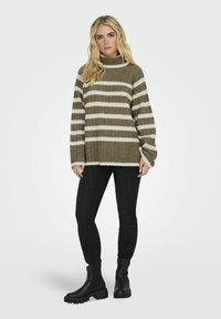 Neulepusero oliivinvihreänä, valkoisilla raidoilla; korkea kaulus, rento malli, pitkät hihat. Yhdistetty mustiin skinny-farkkuihin ja muhkeisiin mustiin saappaisiin.