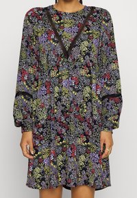 Dlouhá květinová šaty s tmavě modrým pozadím, které obsahují fialové, červené a zelené vzory, a V-výstřih s krajkovými detaily.