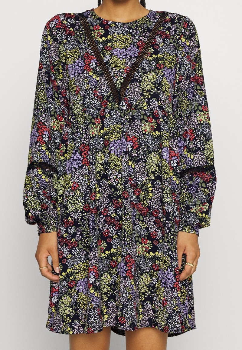 Dlouhá květinová šaty s tmavě modrým pozadím, které obsahují fialové, červené a zelené vzory, a V-výstřih s krajkovými detaily.