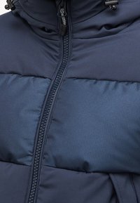 Tmavě modrá prošívaná bunda s polovičním zipem, strukturovanými panely a stahovací kapucí. Hladká látka s kontrastními matnými a žebrovanými sekcemi.