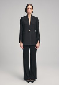 Blazer nero su misura con scollatura a V profonda e bottone dorato, abbinato a pantaloni neri a gamba larga. Tessuto liscio e silhouette strutturata.