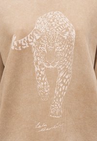 Μπεζ φούτερ με εκτύπωση λευκής λεοπάρδαλης σε στάση τρεξίματος, με λεπτομερείς κηλίδες και υπογραφή της μάρκας με καλλιγραφικά γράμματα από κάτω.