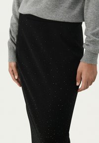 Schwarzer Rock mit einer taillierten Silhouette, verziert mit kleinen, glänzenden Punkten. Weicher Stoff und eine glatte Textur, kombiniert mit einem grauen Pullover.