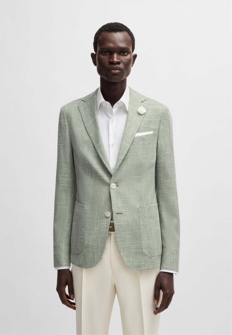 Blazer en lin vert clair avec un motif à carreaux subtil, revers crantés, poches avant et un mouchoir blanc. Associé à un pantalon crème.