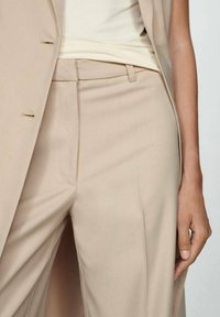 Pantalons beige sur mesure avec une texture lisse, présentant un devant plat, des poches latérales et des détails de couture minimalistes.