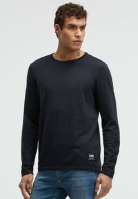 Schwarzes Langarmshirt aus weichem Stoff, mit Rundhalsausschnitt und einem kleinen Logo-Aufnäher am Saum. Kombiniert mit blauen Jeans.