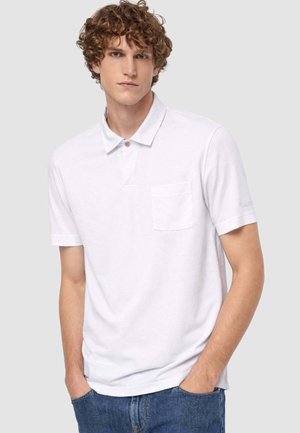 Giovane uomo con capelli ricci che indossa una polo bianca a maniche corte con taschino sul petto e jeans blu, in piedi con le mani nelle tasche.