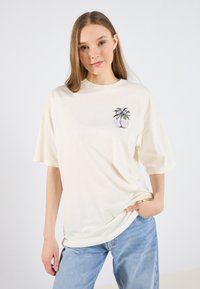 Camiseta de gran tamaño color crema, hecha de algodón, con un gráfico de una palmera negra y la palabra "Aloha" en púrpura. Mangas cortas, corte relajado.