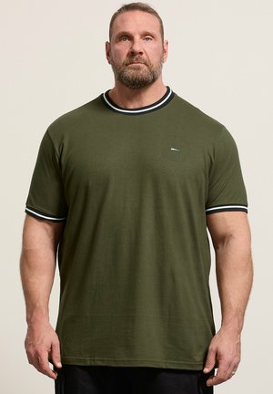 Mand med skæg iført olivengrøn kortærmet t-shirt med sort og hvidstripet krave og ærmekanter, står mod en ensfarvet baggrund.