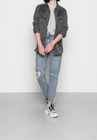 Donkergrijze button-up shirt over een lichtgrijze trui, gecombineerd met versleten lichtblauwe jeans en chunky witte sneakers.