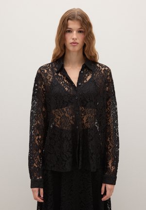 Giovane donna con capelli ricci che indossa una blusa nera trasparente in pizzo con bottoni sopra un reggiseno nero abbinato, in piedi davanti a uno sfondo chiaro e neutro.