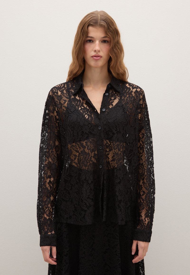 Giovane donna con capelli ricci che indossa una blusa nera trasparente in pizzo con bottoni sopra un reggiseno nero abbinato, in piedi davanti a uno sfondo chiaro e neutro.