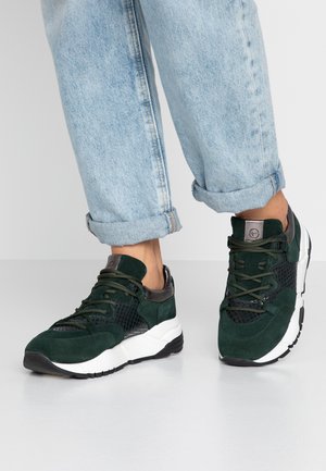 Person wearing rolled-up light blue jeans and dark green suede sneakers with white soles standing on a plain floor.