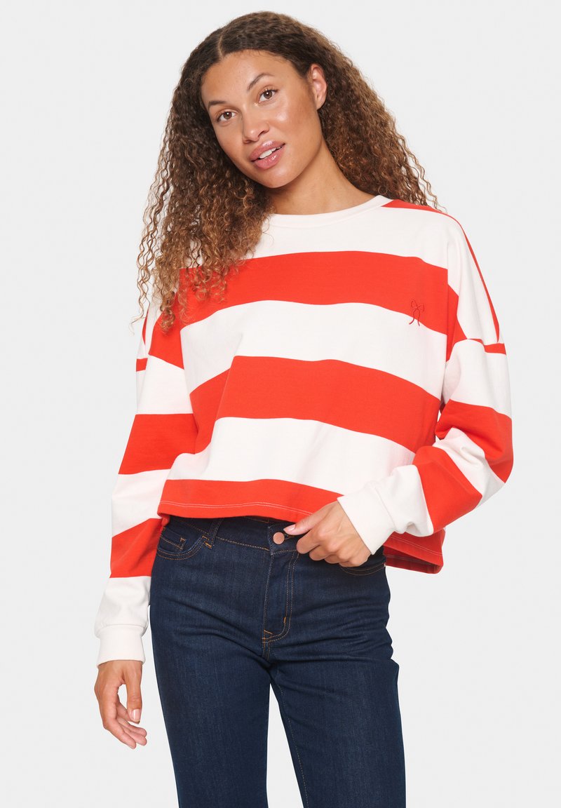 Felpa cropped a righe rosse e bianche con maniche lunghe, caratterizzata da un dettaglio ricamato sul petto, abbinata a jeans blu scuro.