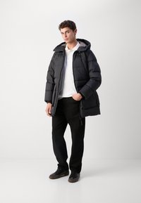 Schwarze Daunenjacke mit Kapuze, gestepptem Design und Reißverschluss vorne, getragen über einem weißen Poloshirt und schwarzen Hosen, mit dunklen Sneakers.